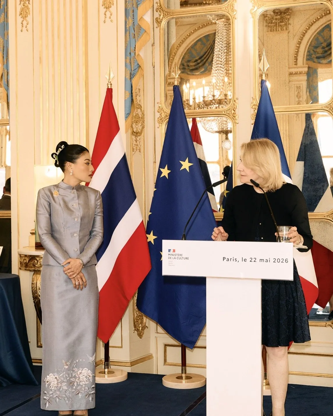 A Thai princess in a grey silk dress stands gracefully beside a French official speaking at a podium bearing the text "Paris, le 22 mai 2026."