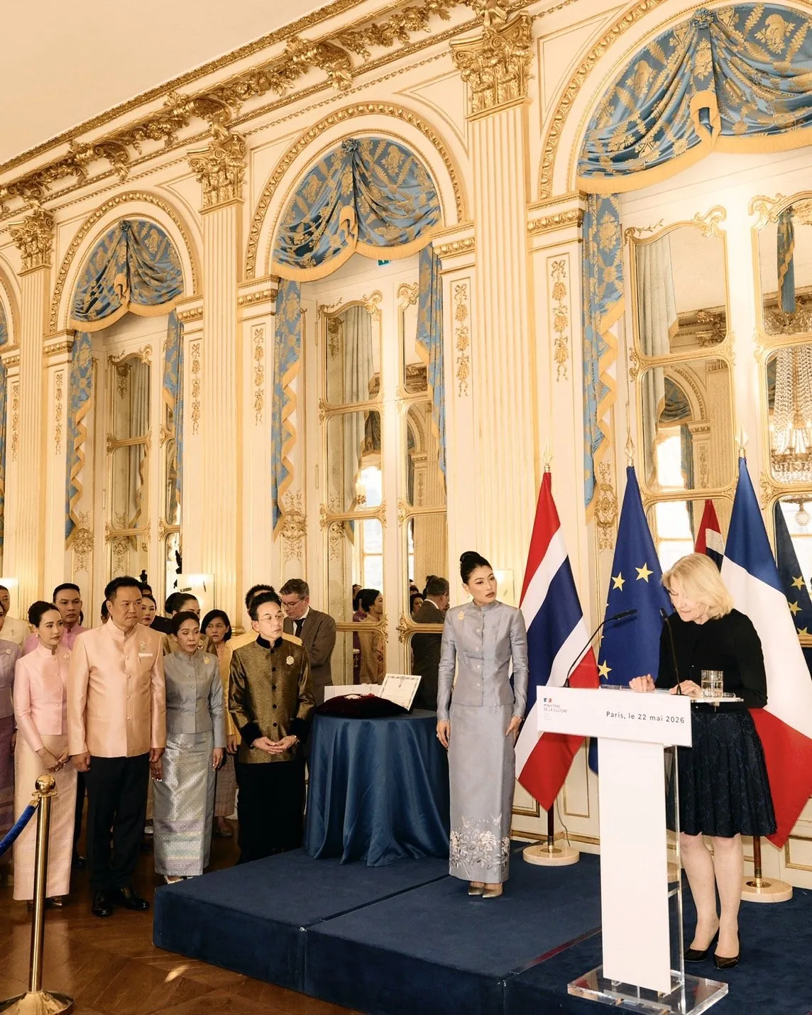 A wide view of the ornate golden hall. Attendees in formal attire watch as the French official stands at the podium with the princess.