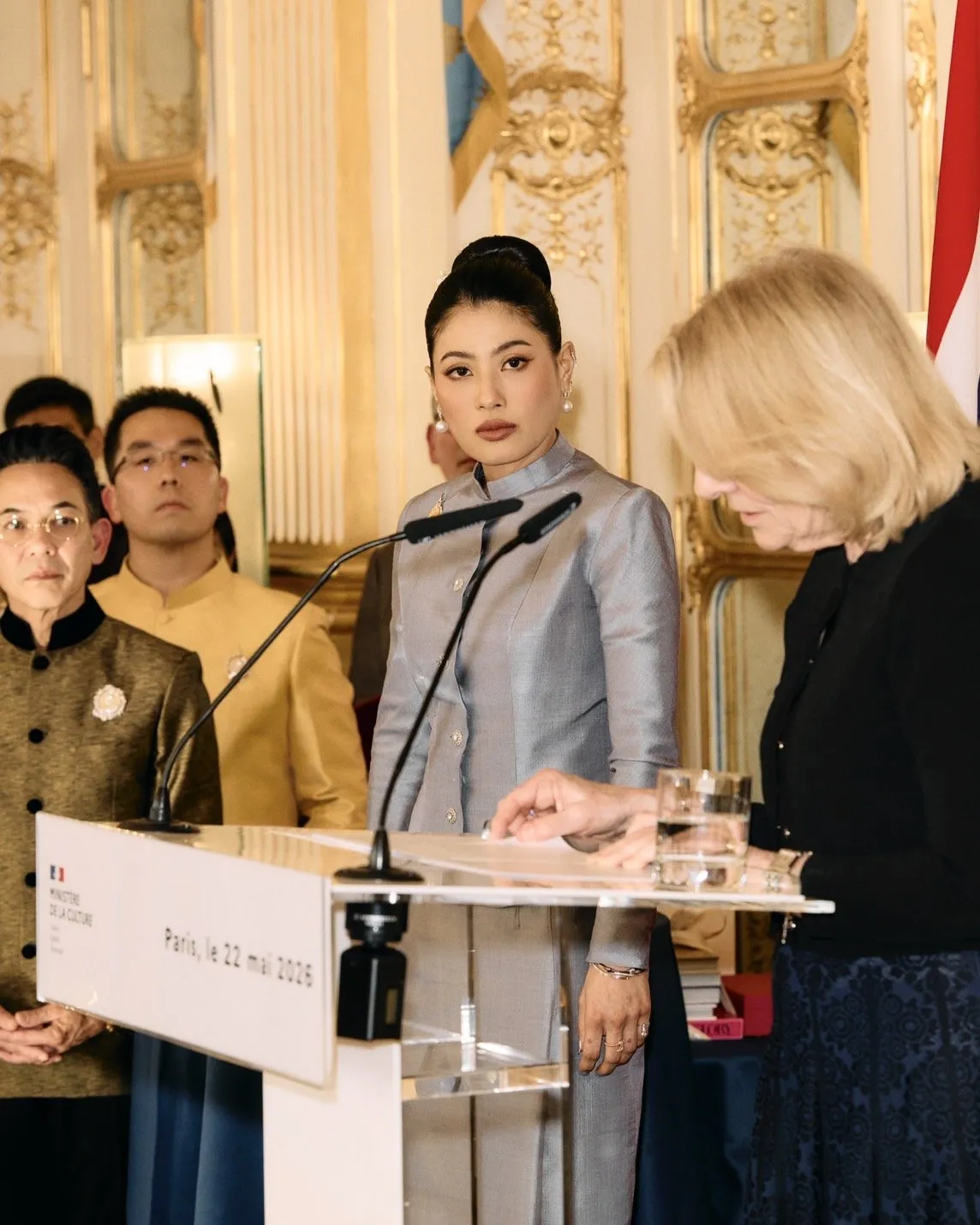 A close-up of the Thai princess looking toward the camera while the French official reviews papers at the podium.
