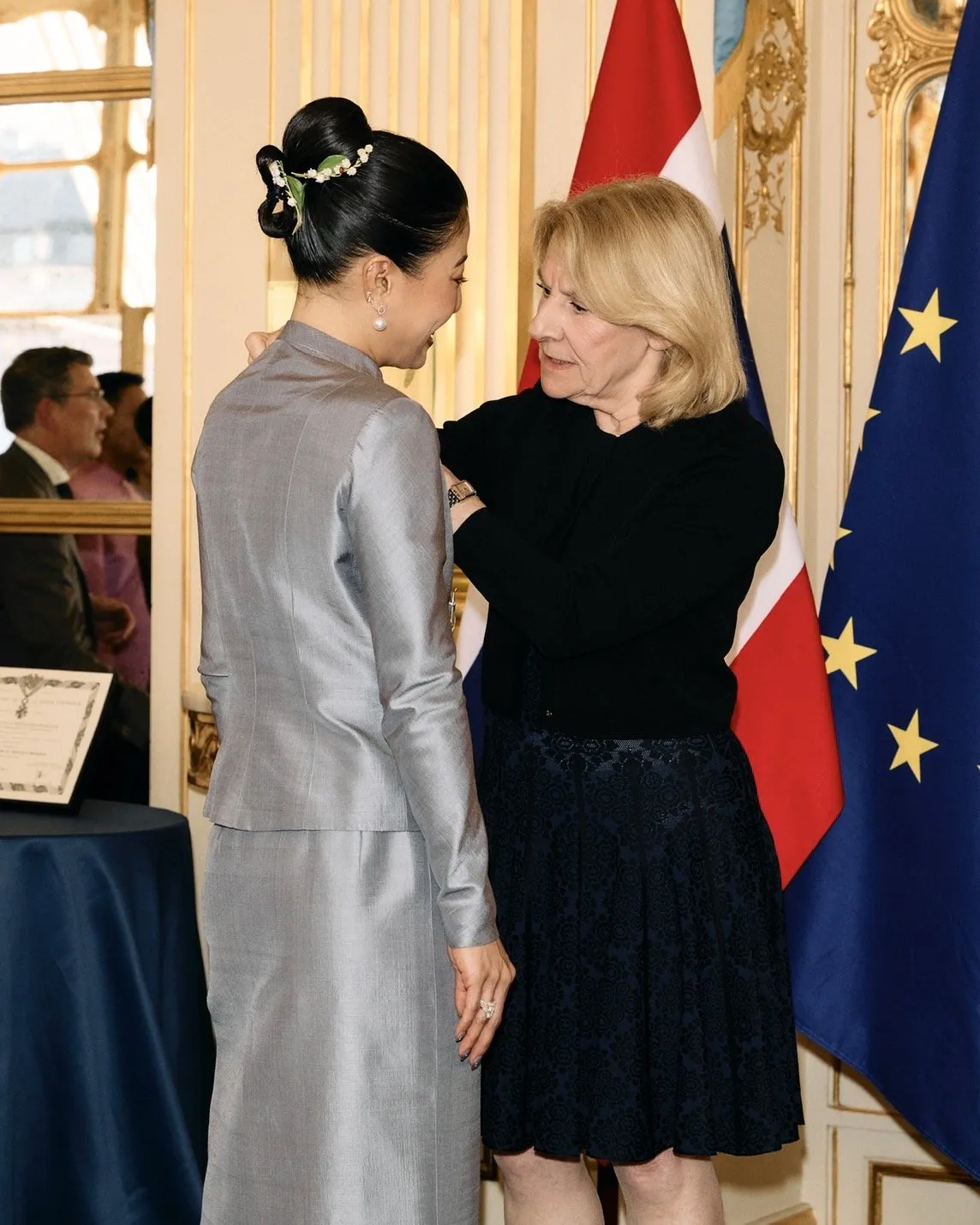 The French official pins the medal onto the princess's shoulder against the backdrop of the Thai and European Union flags.
