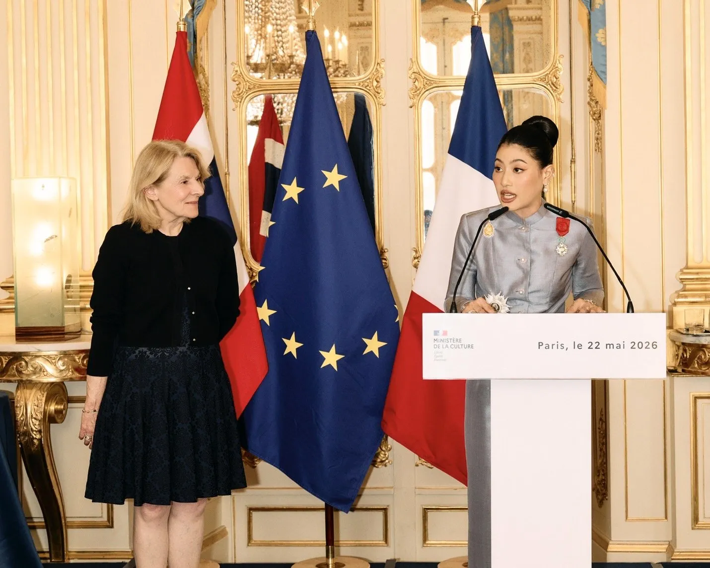 The Thai princess speaks into the podium microphones, wearing the newly pinned medal, while the French official looks on with a smile.