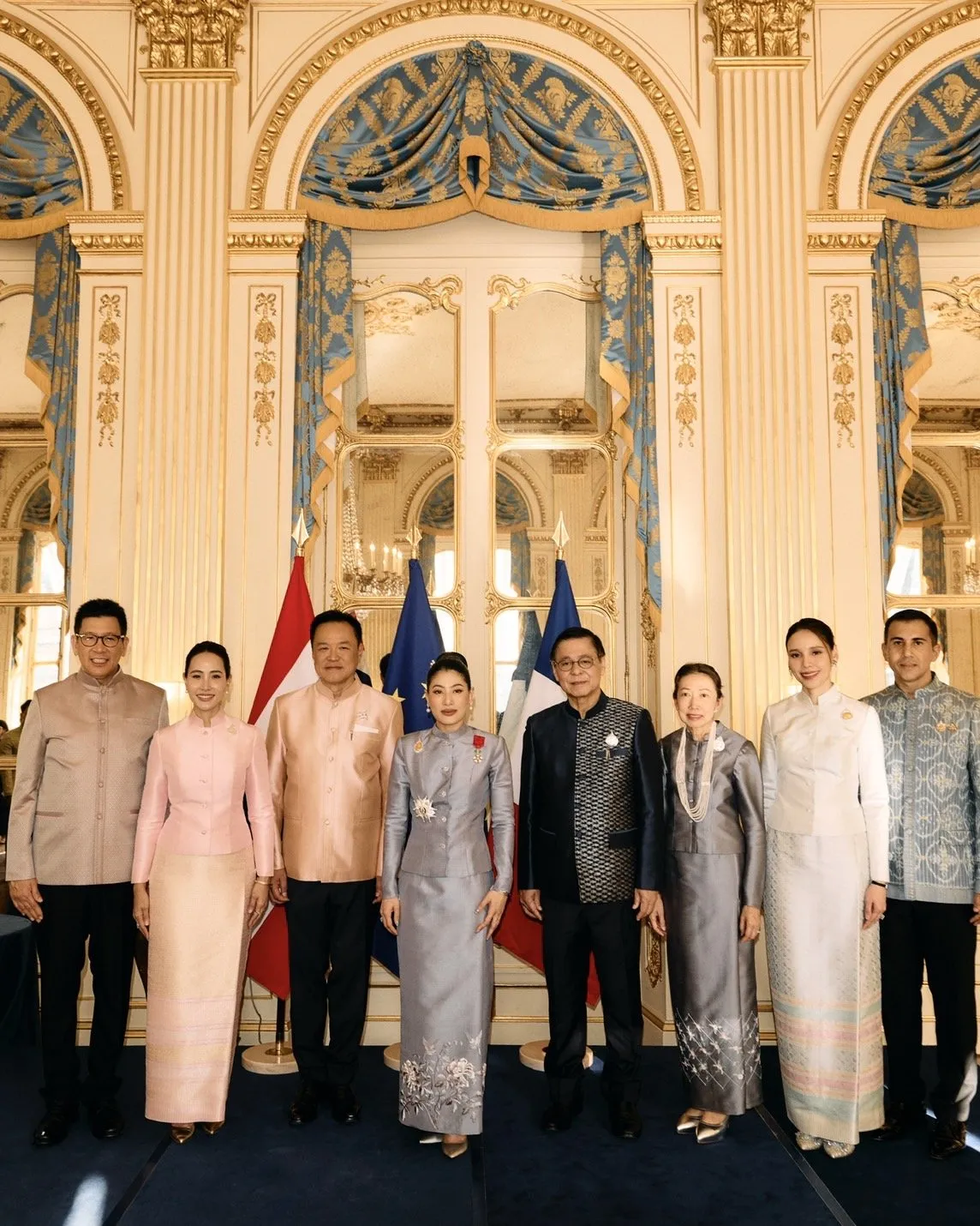 The princess stands with a group of Thai officials in formal attire, posing together inside the ornate, gold-decorated French hall.