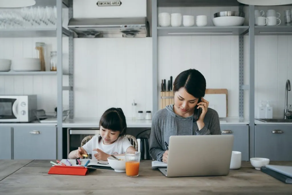 A mother talking on the phone with a child coloring a book next to her
