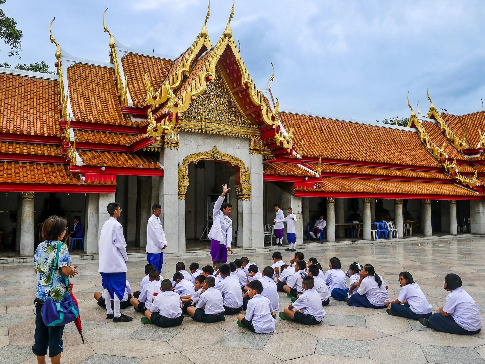 a group of students sitting on the ground being supervised by a group of adult. they are in a temple.