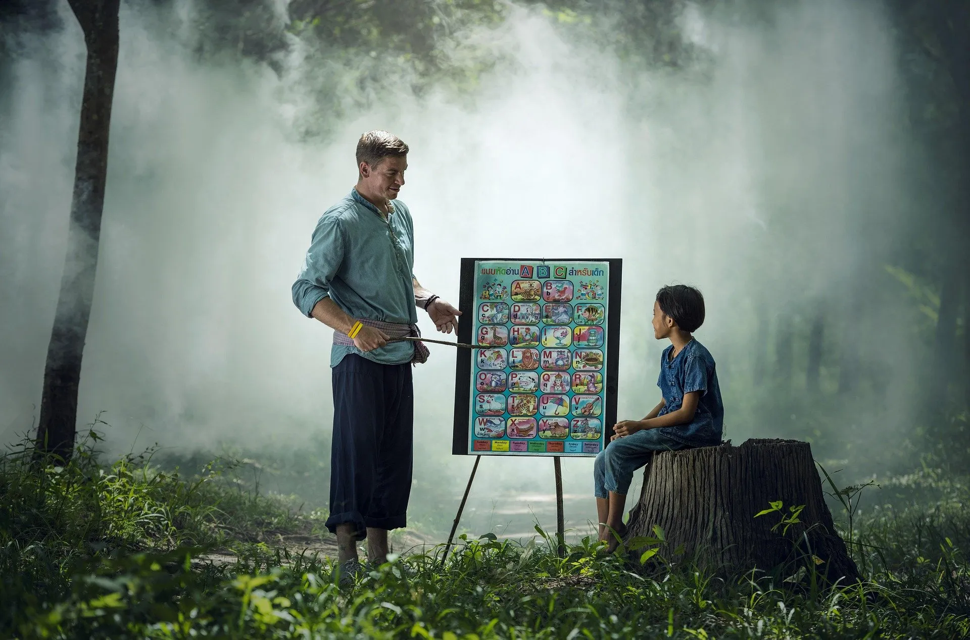 a Caucasian male teacher giving a lecture to a lone Thai boy in a brightly lit forest. the boy is sitting on a stumb.