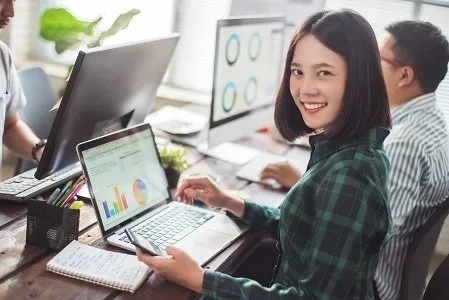 A woman happily working and smiling at the camera with her laptop and cell phone out.