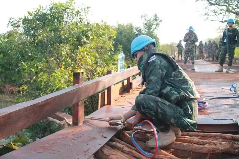 Thai peacekeepers in military fatigues and blue U.N. helmets, reparing a wooden bridge over a gorge.