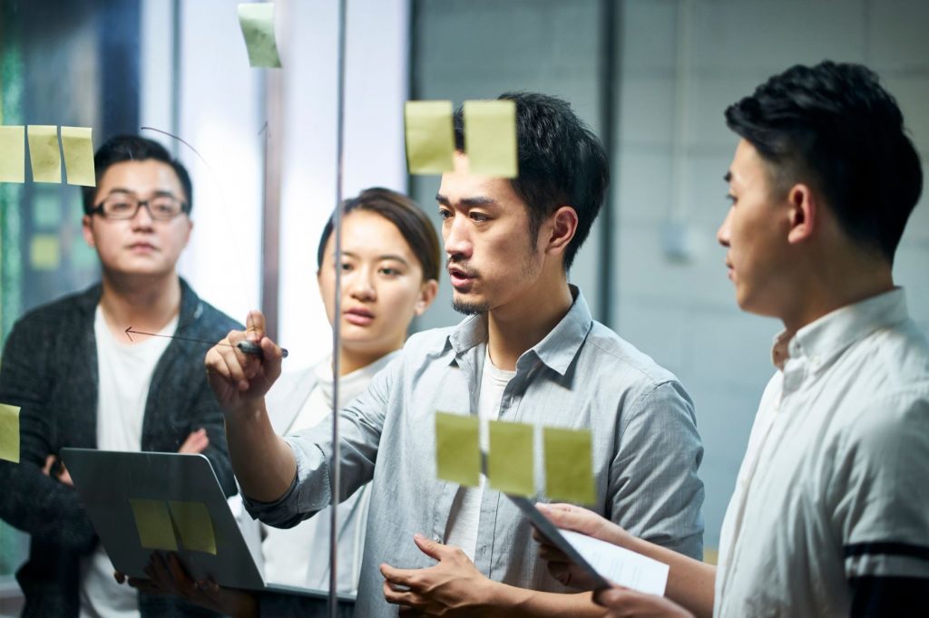 Man in startup board meeting presenting idea with sticky notes and writing on glass panel.