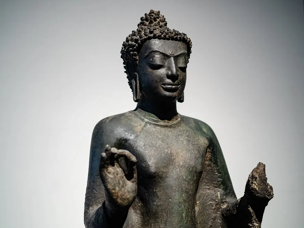 An aged bronze-casted standing buddha in a blessing position on display at the National Museum Bangkok
