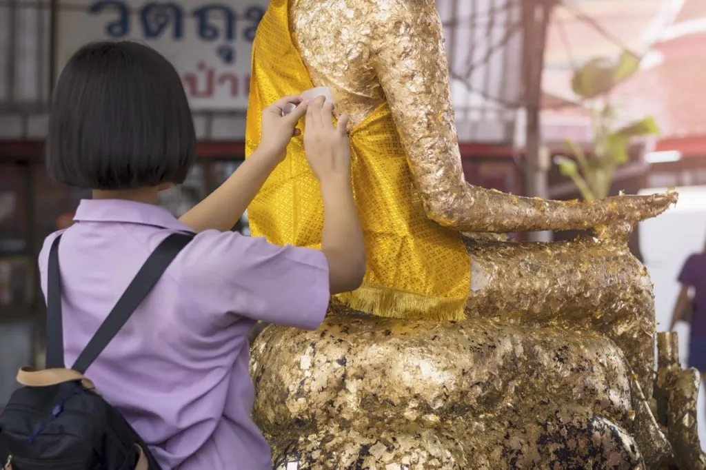 A Thai student applying a gold leaf at the back of a Buddha statue