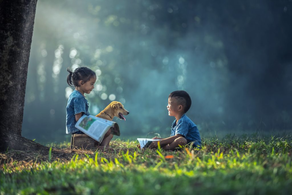 Two kids and a dog sitting facing each other on the grass