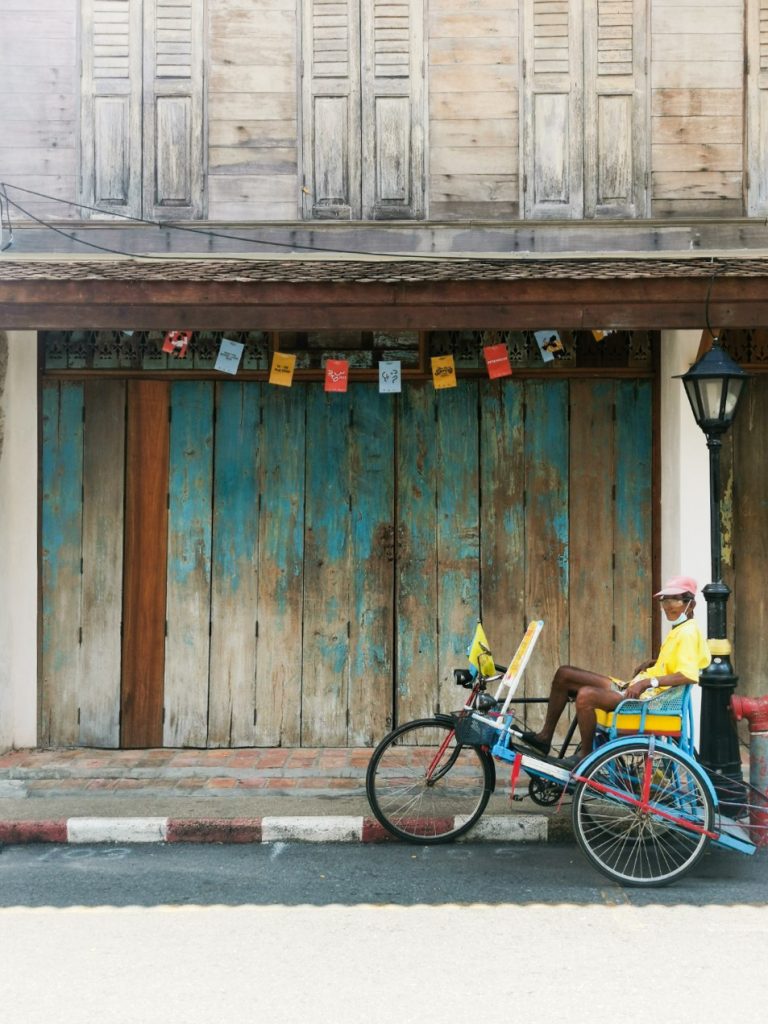 An old man sitting on a tricycle taxi in front of an old building with Pakk Taii Design Week posters