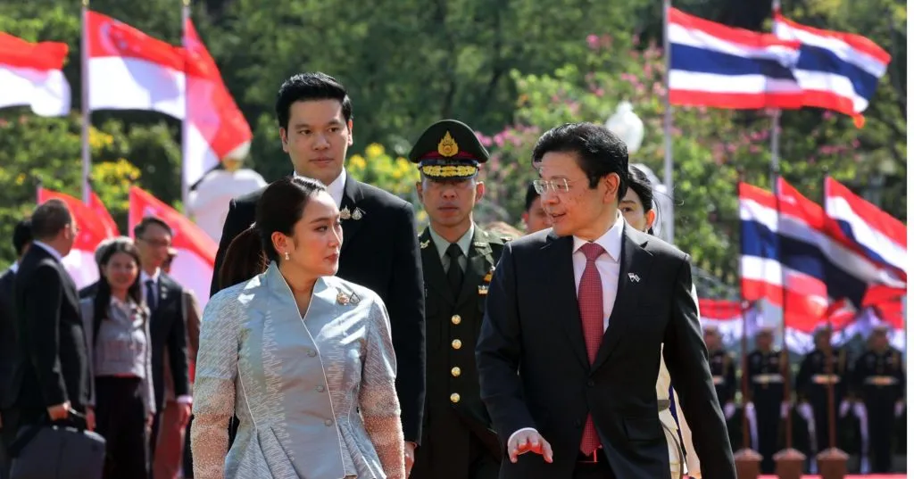 female in blue silk talking to a male in deep blue suit and red necktie with two male trailing behind them. guards standing in the background. flags of Singapore and Thailand are also placed in the background.