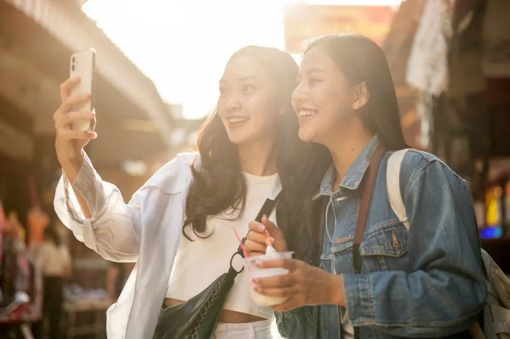 Two pretty, joyful young Asian female tourists enjoy exploring street food at a market in Thailand on a sunny day, taking a selfie together with a smartphone while walking along the street.