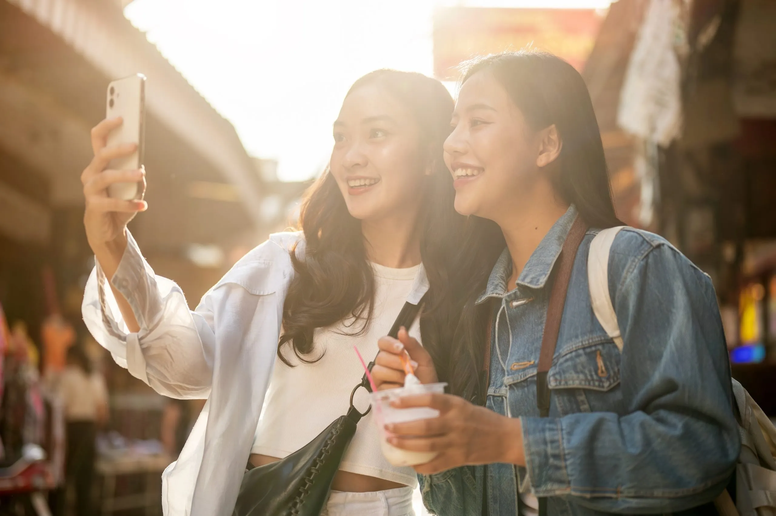Two pretty, joyful young Asian female tourists enjoy exploring street food at a market in Thailand on a sunny day, taking a selfie together with a smartphone while walking along the street.