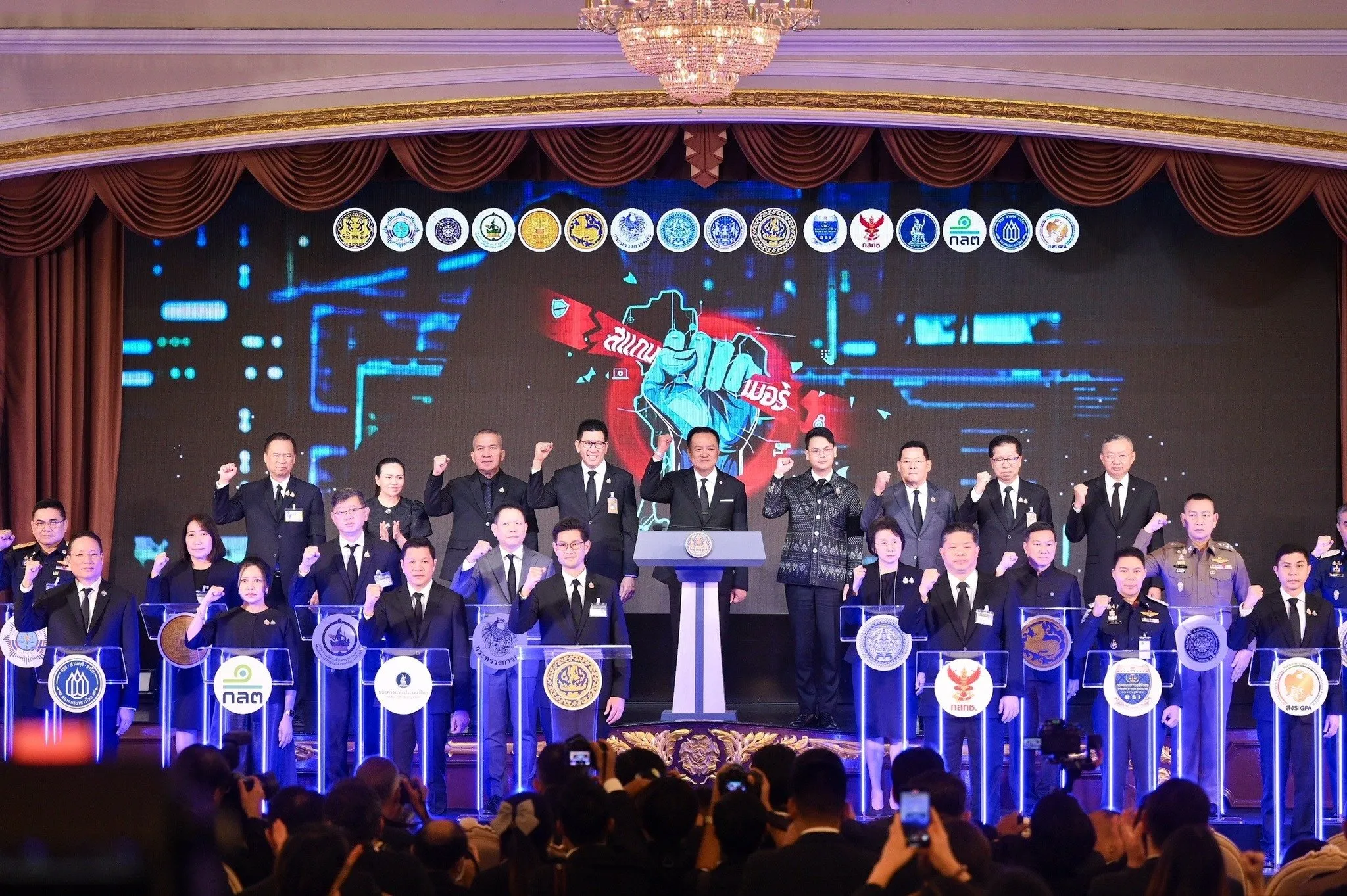 The Prime Minister and leaders from 15 major government agencies stand together on a brightly lit stage, each at their own podium. They raise their fists in a show of unity, wearing dark formal suits. Behind them is a large digital screen displaying the image of a clenched blue fist splitting through a graphic of Thailand, with the words “Scammers” in bold red Thai script, and the emblems of all participating agencies arrayed along the top.