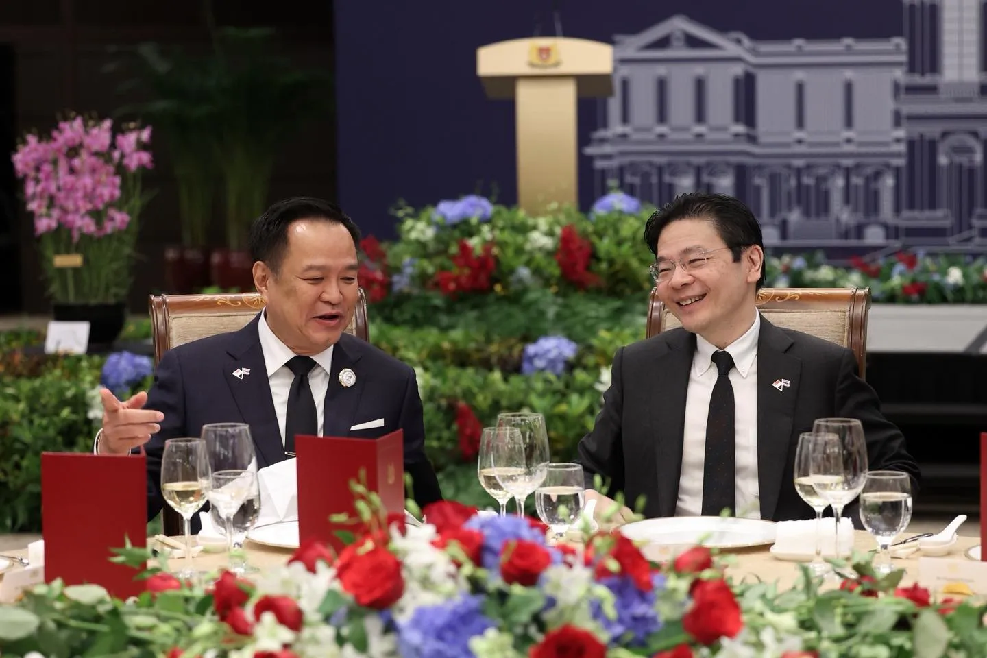 Prime Minister Anutin Charnvirakul of Thailand and Prime Minister Lawrence Wong of Singapore in dark suits are seated side by side at a formal dinner table, deep in conversation. The table is set with wine glasses, water glasses, and red menus among fresh arrangements of red, white, and purple flowers.