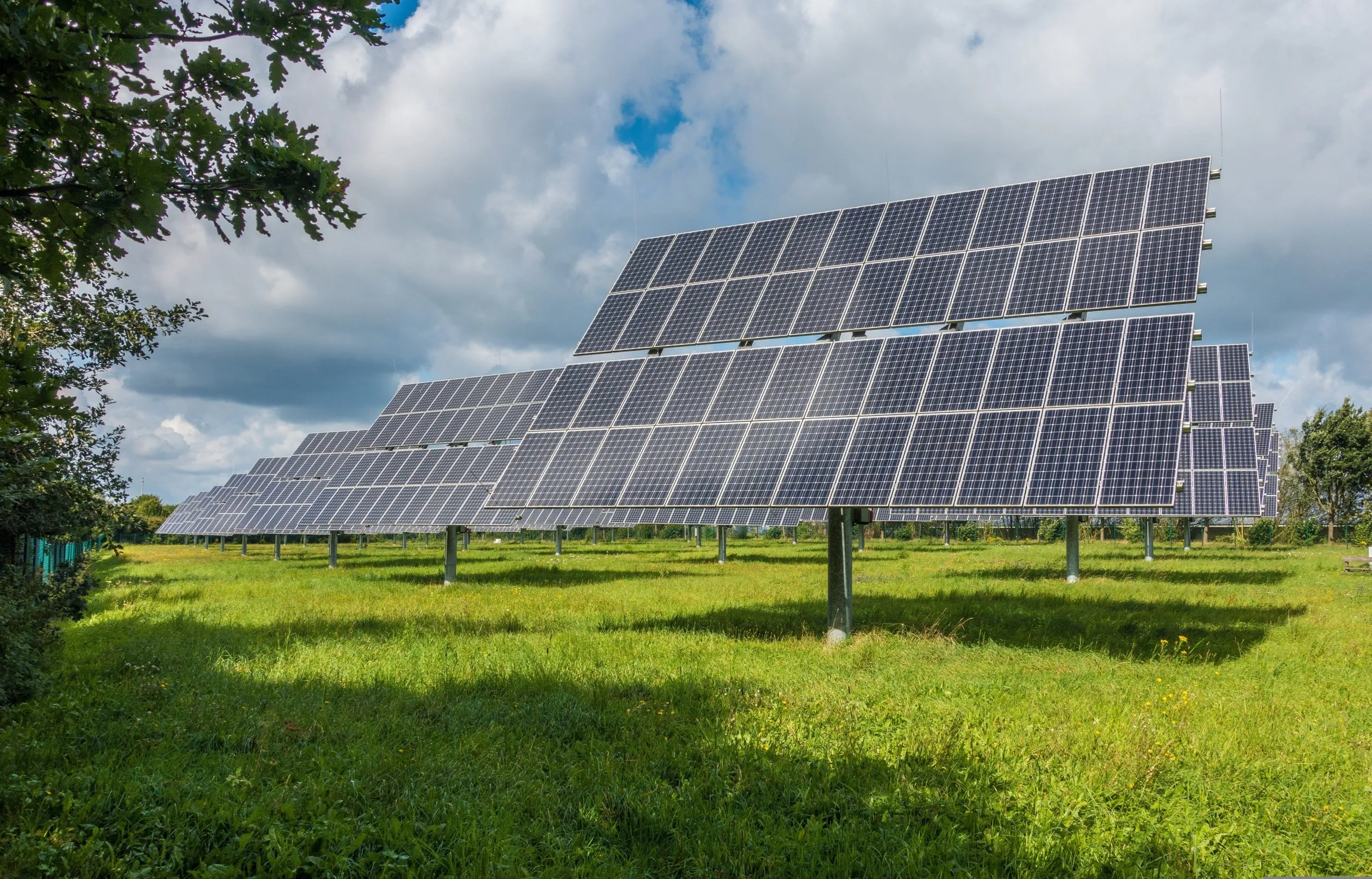 Rows of large, tilted solar panels standing on grassy green fields under a partly cloudy blue sky.