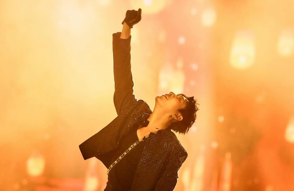 A lone male singer standing, looking up, and raising his right fist. He is performed in front of a bright orange background.