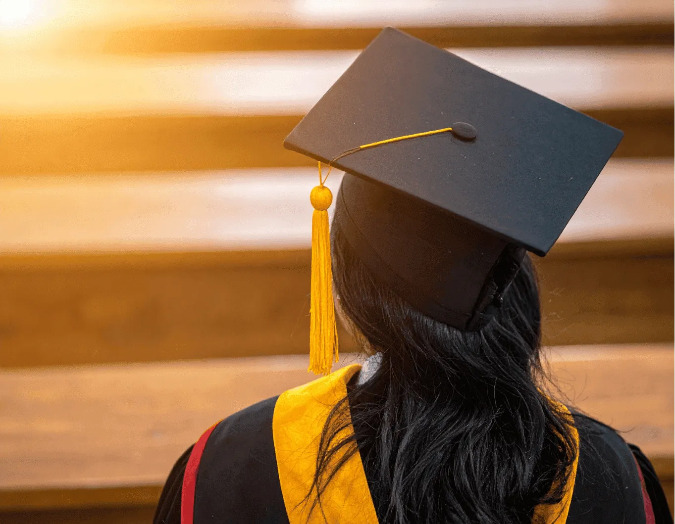 A female scholar in her gown dress and hat turning her back to the camera with bright orange light in the background