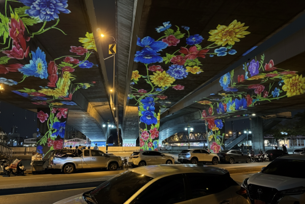 Vibrant blue and yellow flower projections cover the underside of a dark concrete highway overpass, illuminated above parked cars at night.