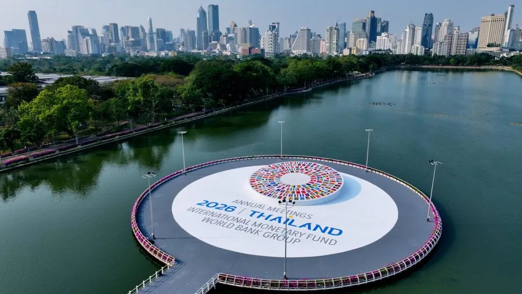 This drone shot captures a white circular platform on a lake, displaying 2026 meeting text and a centerpiece covered in world flags.
