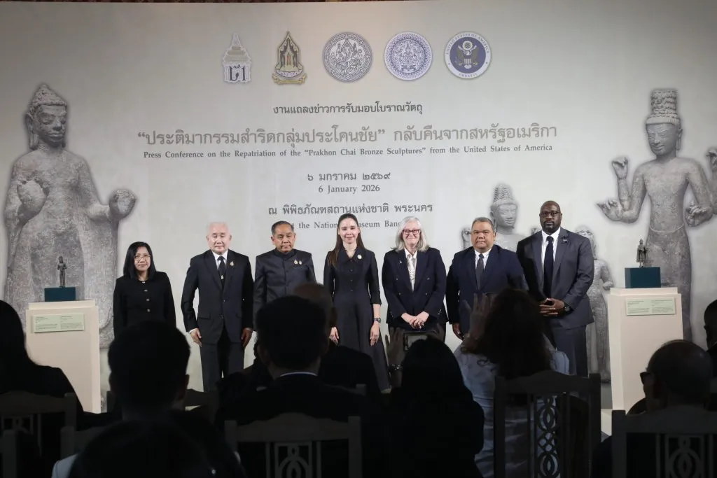 Seven officials stand on a stage with a backdrop of text and sculptures, addressing a seated audience at a press conference.