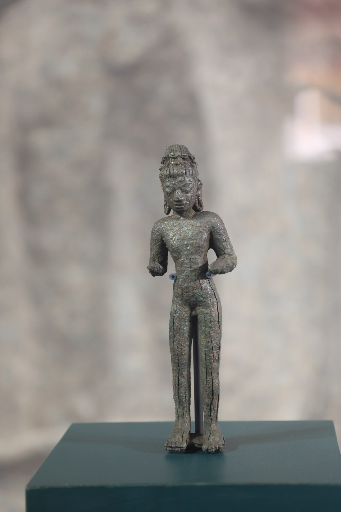 A slender, weathered ancient bronze deity stands upright. Its rough, greenish-brown surface features a tall bun, four arms, and a simple, textured loincloth.
