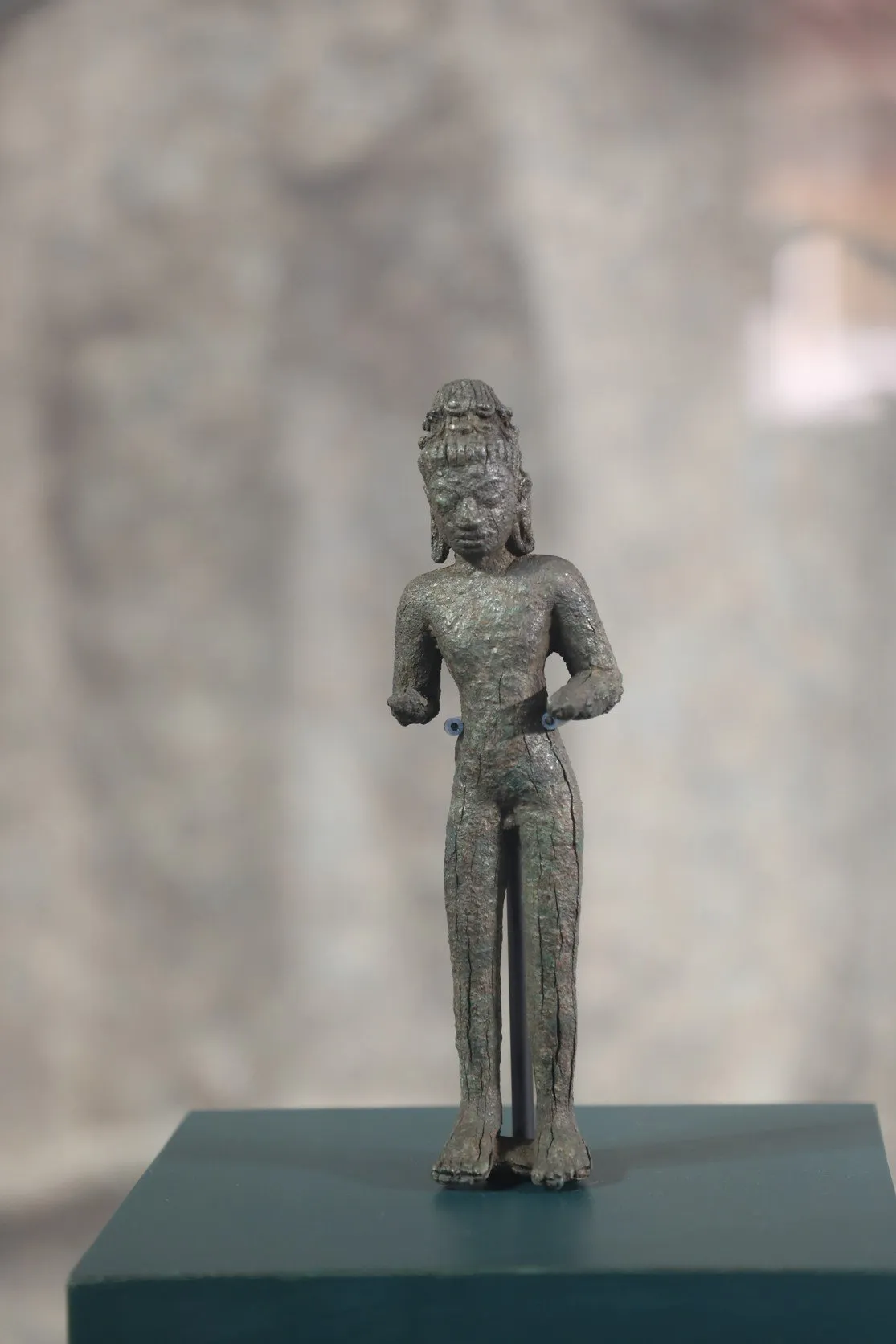 A slender, weathered ancient bronze deity stands upright. Its rough, greenish-brown surface features a tall bun, four arms, and a simple, textured loincloth.