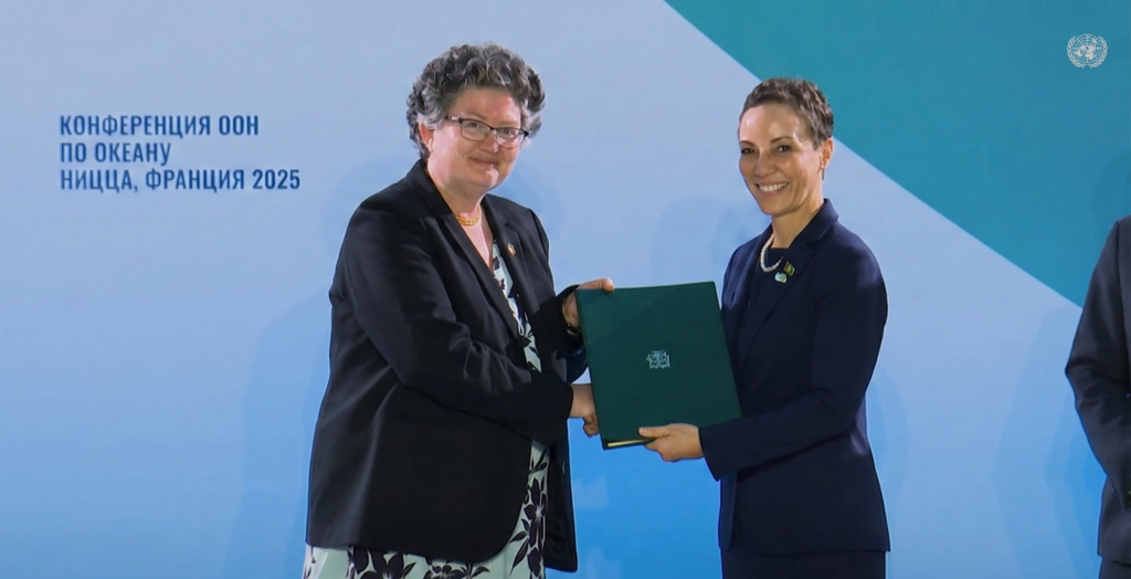 Two smiling women in business jackets shake hands and hold a green folder at a table. They are in front of a light blue wall with text and a UN logo.