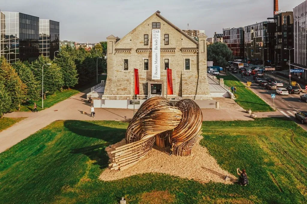 Curved wooden planks form a large, knotted sculpture on a sunlit lawn. Behind it stands a tall stone building with vibrant red banners.