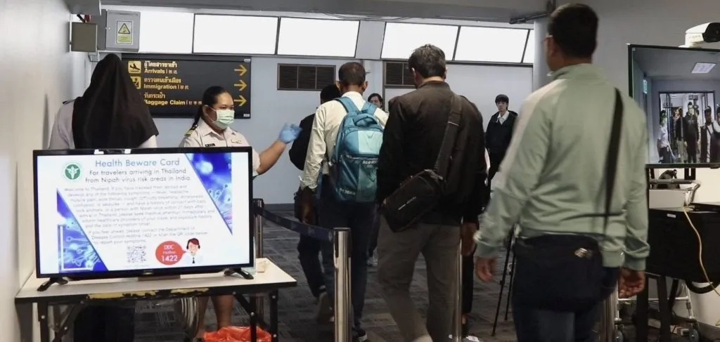 At a Thai airport, a uniformed official wearing a mask gestures toward a digital "Health Beware Card" screen regarding Nipah virus screening.