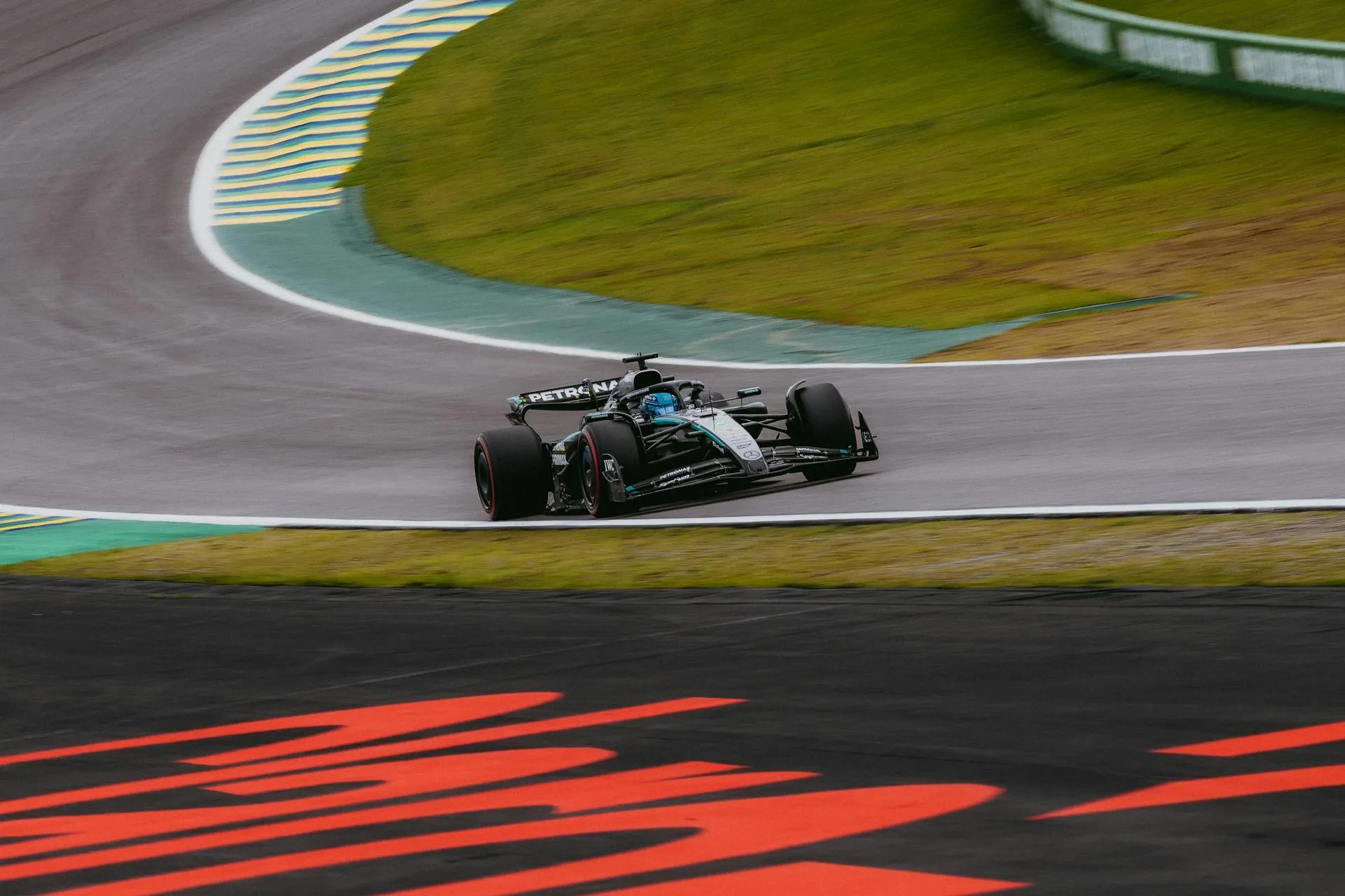 A sleek, black and silver Formula One car speeds around a grey racetrack curve, bordered by vibrant green grass and yellow-striped curbs.
