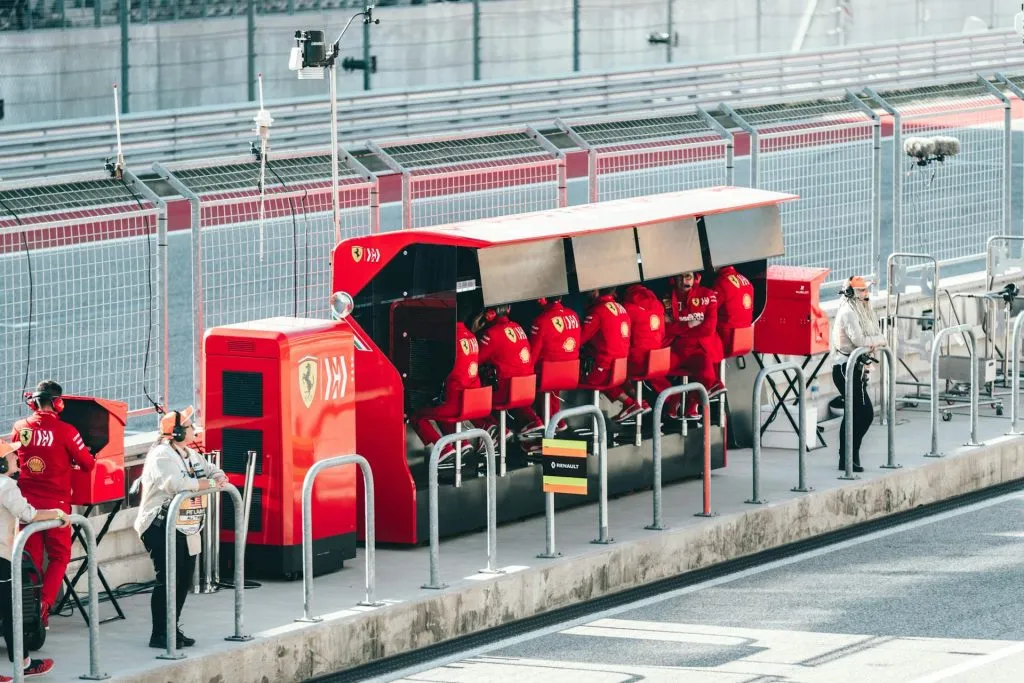 A row of Ferrari team members in bright red uniforms sit at a trackside pit wall station, facing away from the camera.