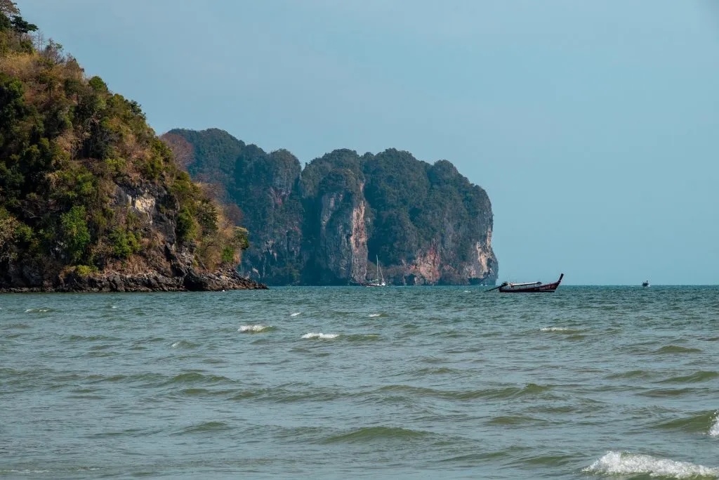 Choppy green waves ripple in the foreground. A traditional longtail boat sails past towering limestone cliffs blanketed in lush greenery, all under a vast, cloudless blue sky.
