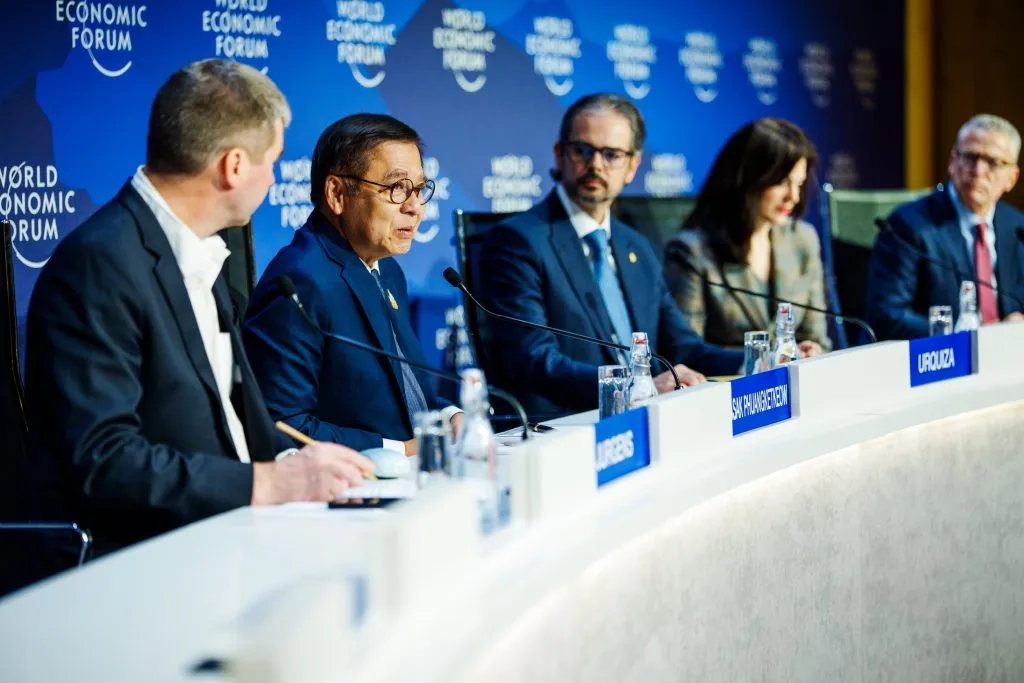 Five professionals sit behind a long white desk during a World Economic Forum panel; an Asian man speaks, backed by a blue wall of logos.