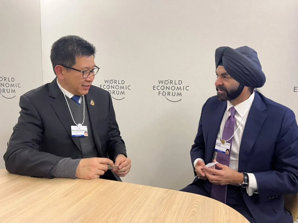 Two men sit at a wooden table in front of a World Economic Forum logo wall, exchanging business cards.