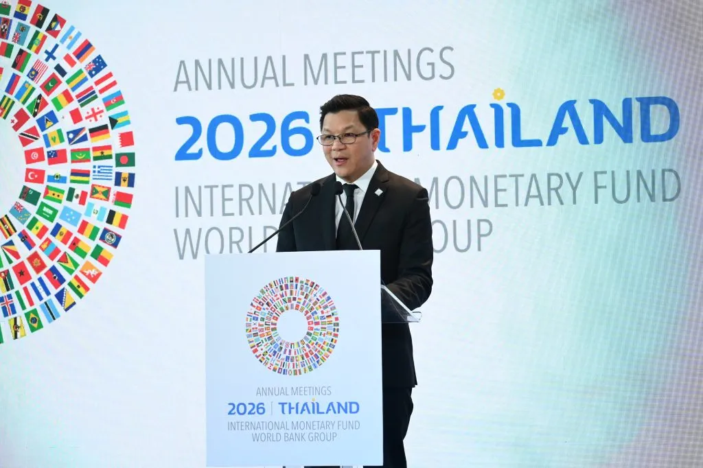Vitai Ratanakorn, wearing glasses and a black suit, speaks behind a podium featuring a circular flag logo for Thailand's 2026 IMF World Bank meetings.