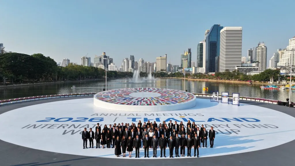 A large group in black suits stands on a white circular stage by a lake, featuring a massive flag logo and city skyline.