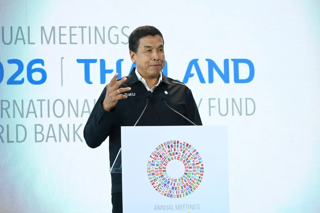 Chadchart Sittipunt, wearing a black jacket, gestures while speaking behind a podium with a colorful circular flag logo for Thailand's 2026 meetings.