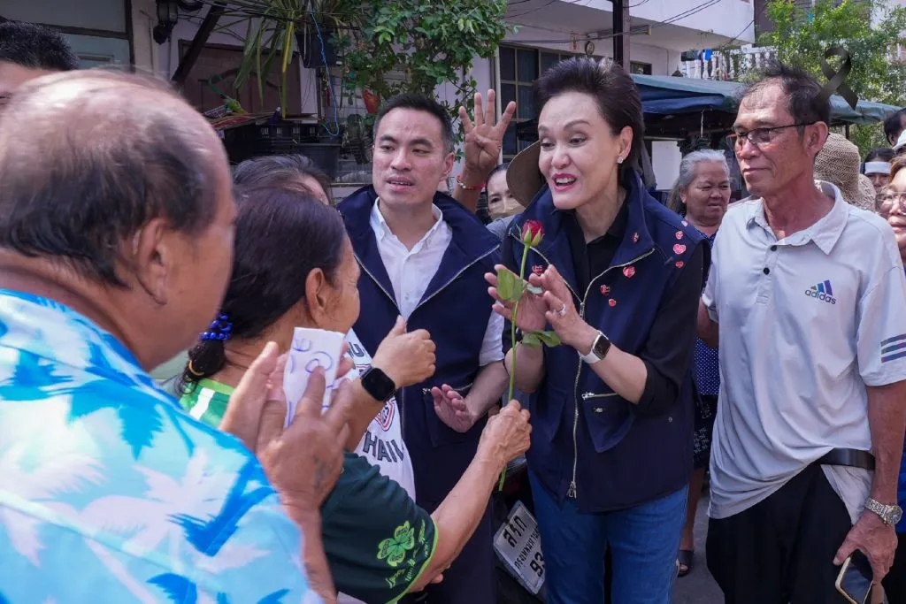 A smiling female politician in a navy vest holds a red rose, engaging with supporters in a crowded street. A black mourning ribbon appears in the top corner.