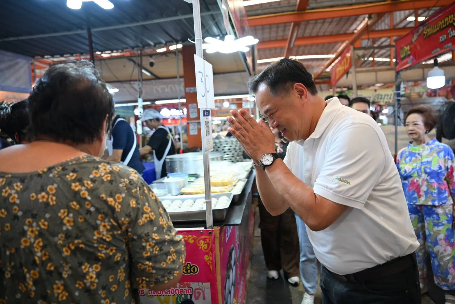 In a busy indoor market, a man in a white polo shirt performs a respectful Thai wai greeting toward a woman standing amidst trays of prepared food.