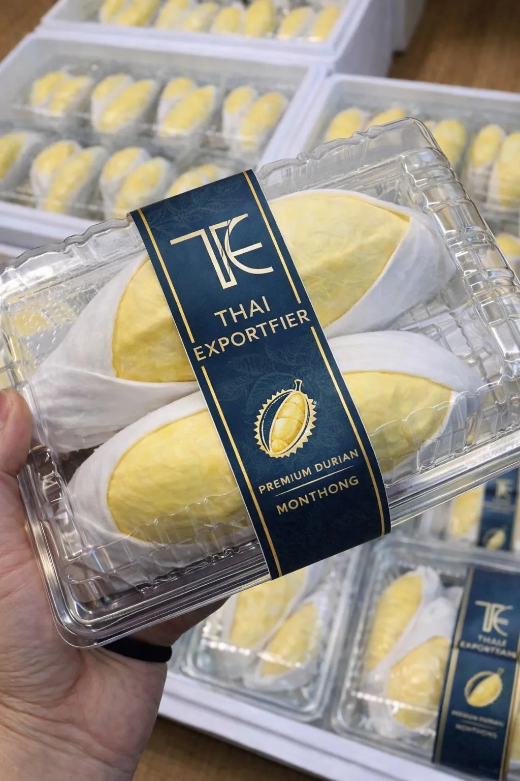A person's hand firmly holds a clear plastic durian package. Many identical packages are neatly arranged in illuminated rows across a surface, defining this indoor commercial setting.