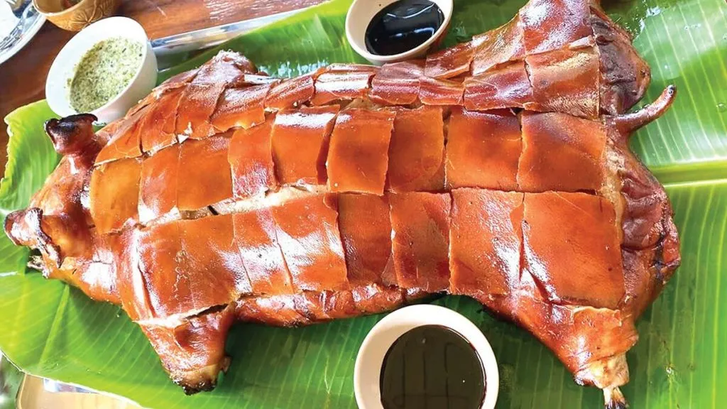 A golden-brown suckling pork, expertly scored, rests on vibrant green banana leaves. Small bowls of dark sauce and green condiment sit on a wooden table, ready for a meal.