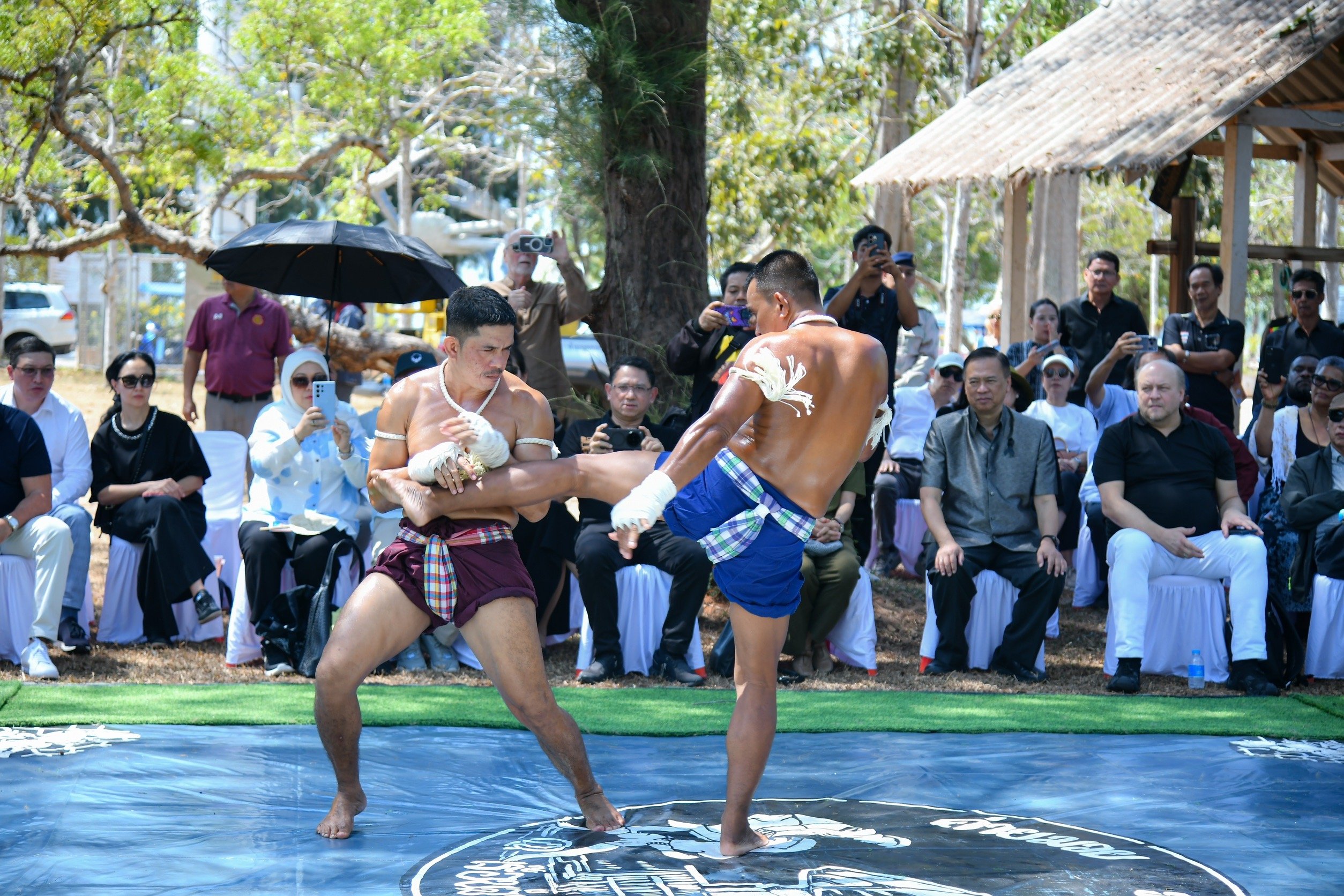 Two shirtless Muay Thai fighters spar on a blue mat, one blocking a kick, before a diverse seated audience. Lush trees and a thatched hut frame the bright outdoor event.