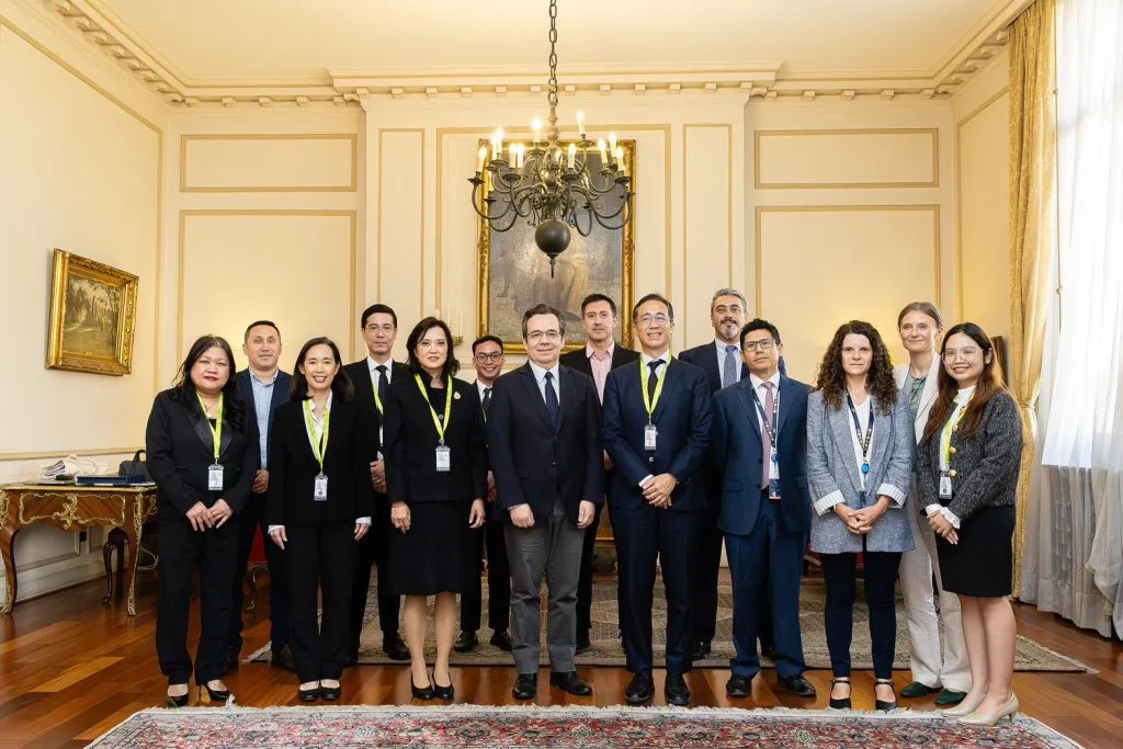 Fourteen formally dressed professionals with lanyards stand smiling on an ornate rug in a grand room with gilded walls, a large chandelier, and a classical painting in the background.