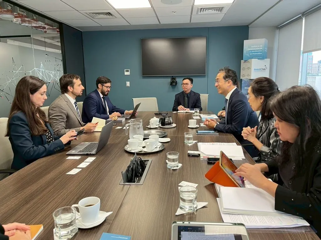 Seven formally dressed business professionals are seated around a large wooden conference table in a modern room, actively conversing and working with laptops.