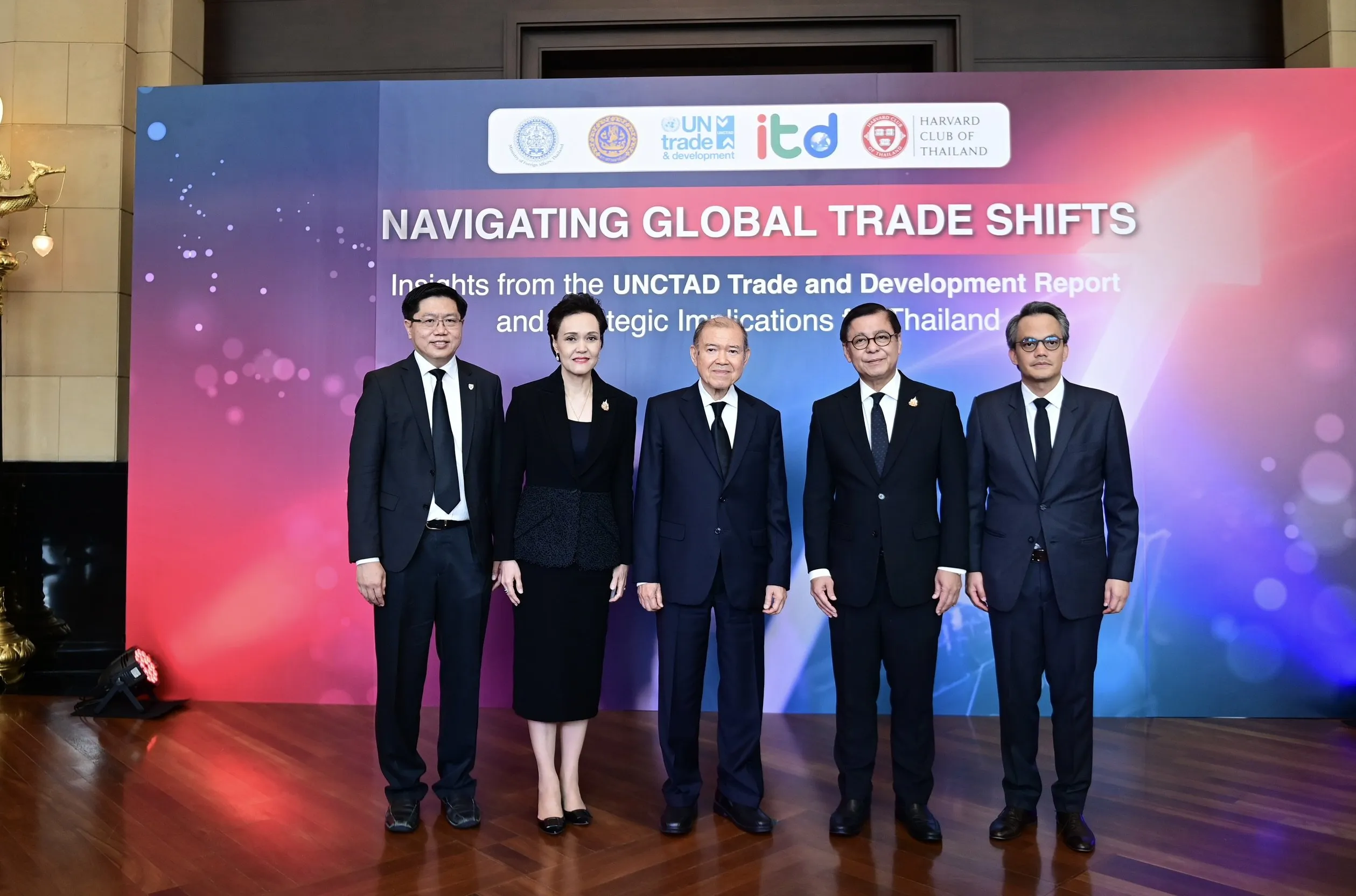 Five formally dressed individuals, one woman and four men, stand smiling on a polished wooden floor in front of a large, colorful conference backdrop.