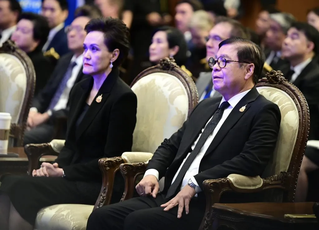A formally dressed man in glasses and a woman sit attentively in ornate chairs at an indoor event. Many other blurred people are seated behind them in the dim room.