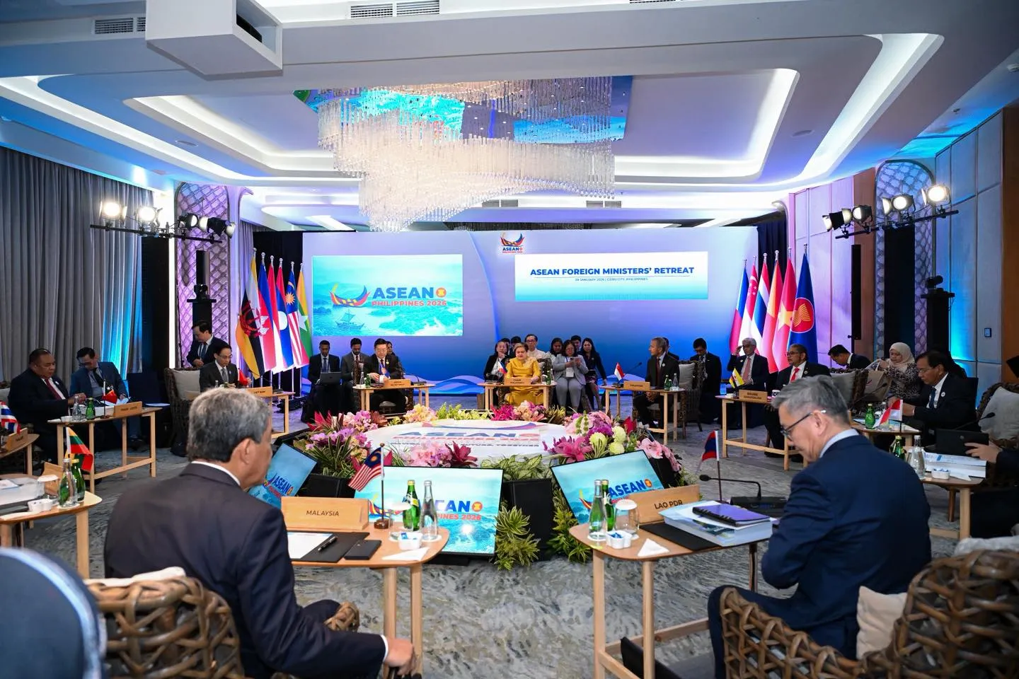 Delegates in formal suits sit in a large circle around a vibrant floral centerpiece in a grand hall, surrounded by national flags for an international diplomatic summit.